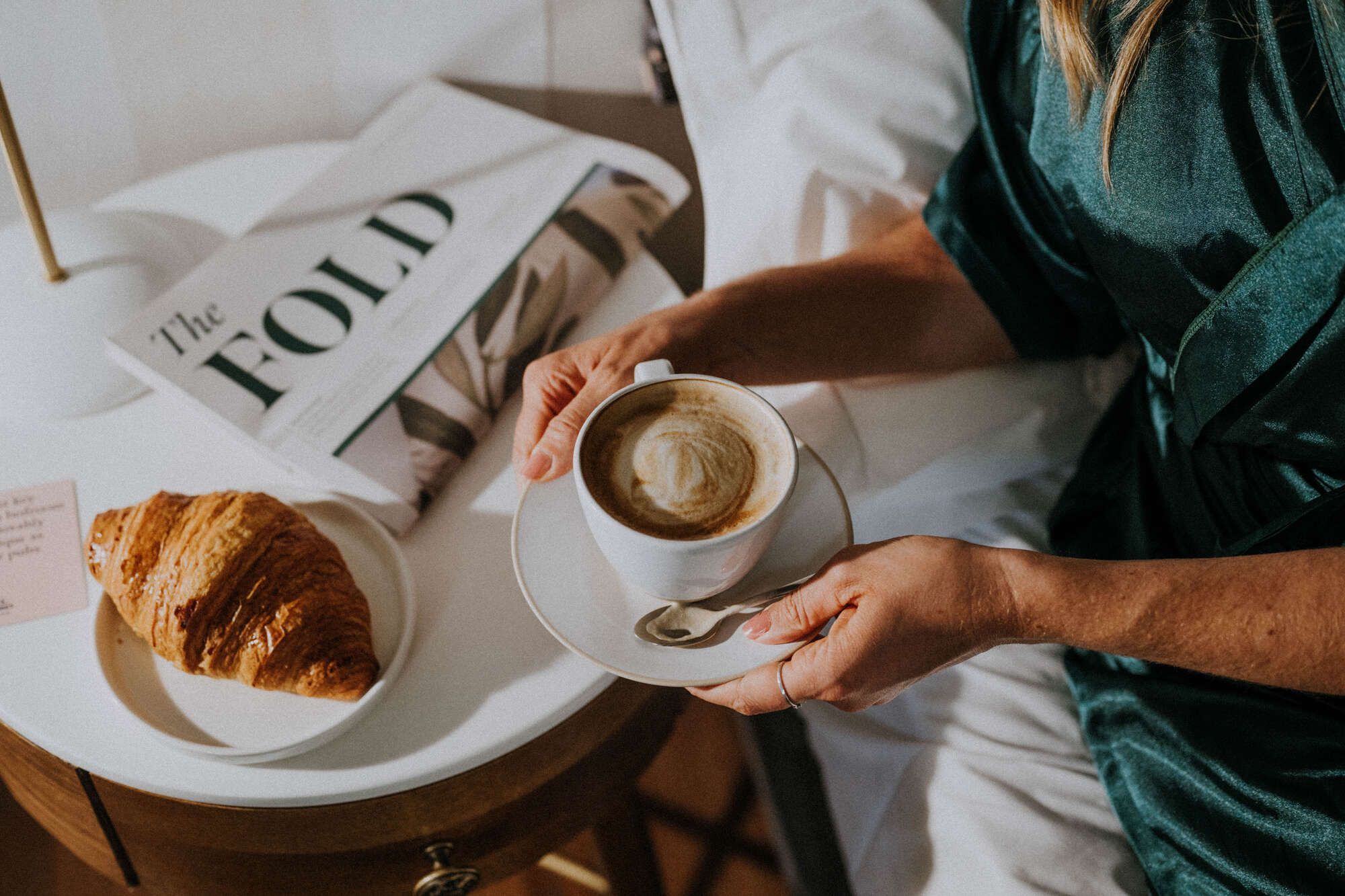 woman holding a coffee with a croissant and an issue of the Fold (Young's in-house newspaper) on the table beside her