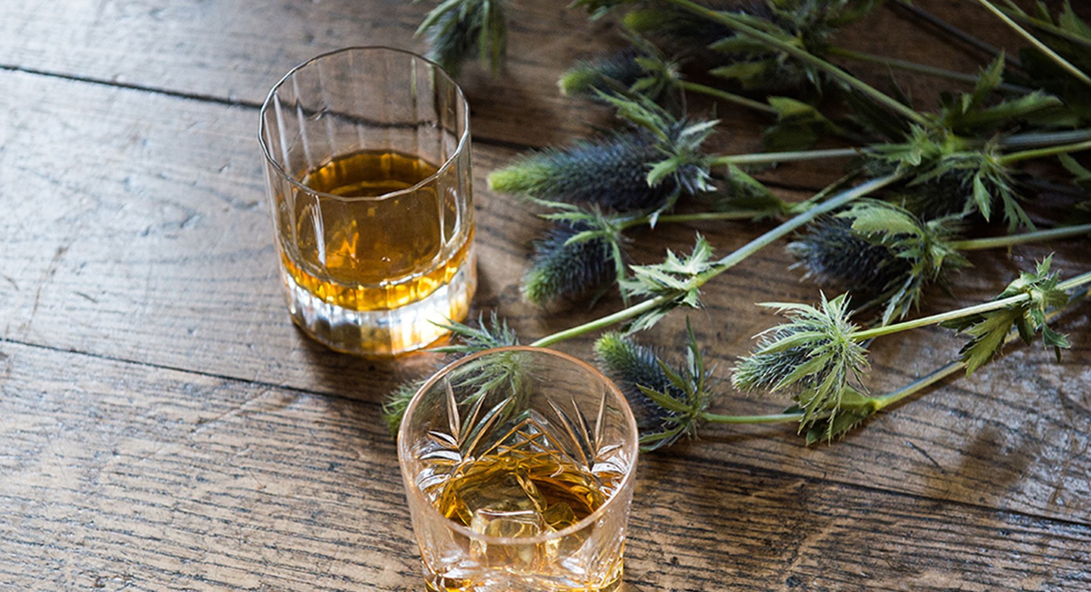 two glasses of whiskey next to flowers on a wooden table